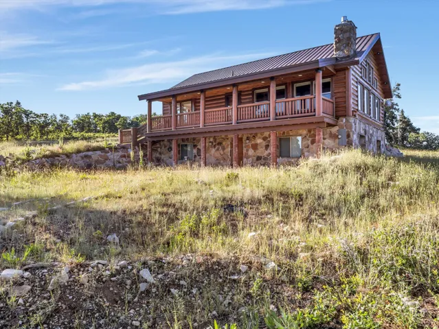 Log cabin exterior featuring a stone foundation, wood siding, a metal roof, an upper-level balcony with wood railings, and a stone chimney