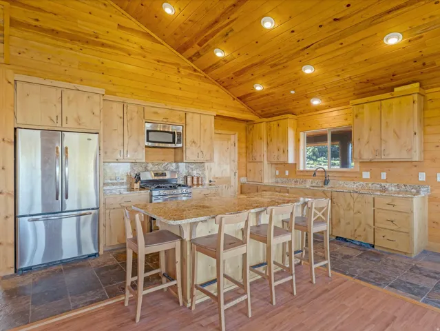 Kitchen featuring a large center island with seating, stainless steel appliances, wood-finish cabinetry, granite countertops, and a vaulted wood-plank ceiling with recessed lighting