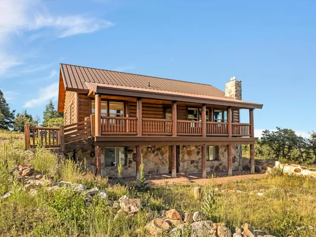 Rustic log-cabin style home featuring a brown metal roof, full-length upper deck with wood railings, stone foundation, and a stone chimney