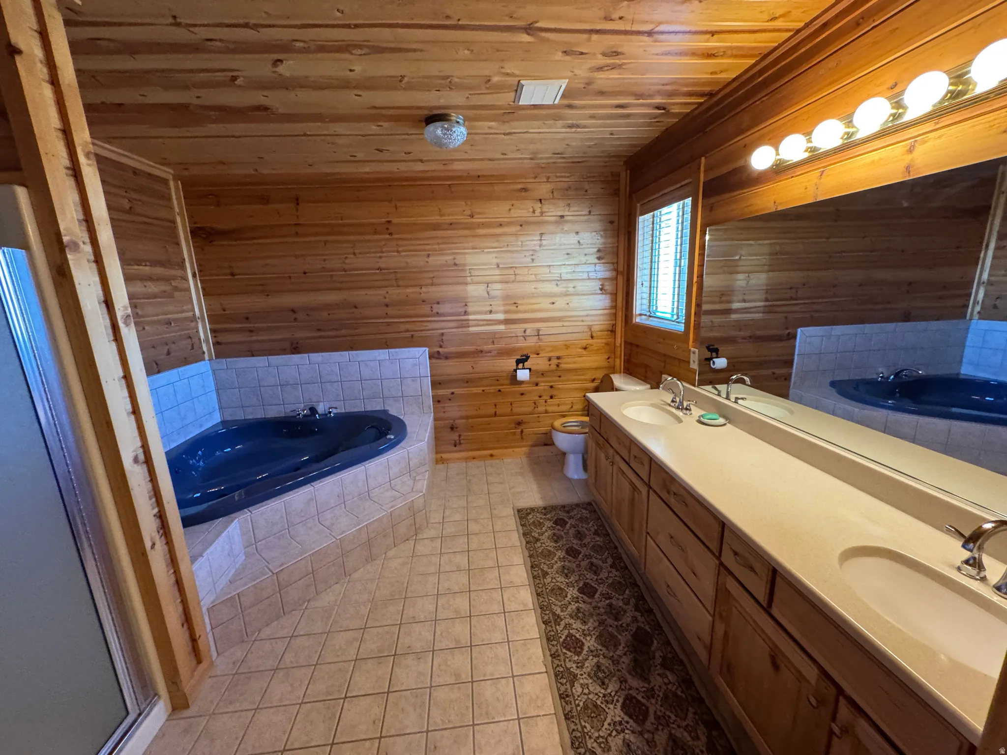 Full bathroom featuring wood paneling, a large corner soaking tub with tiled surround, a double vanity with integrated sinks, and tiled flooring