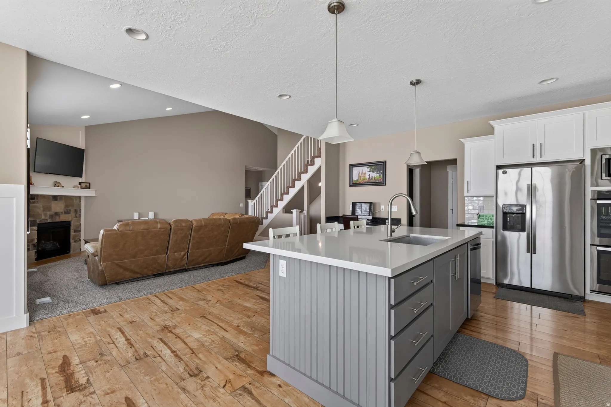Open-concept kitchen and living area featuring a central island with sink, stainless steel appliances, wood-finish flooring, and a stone-faced fireplace