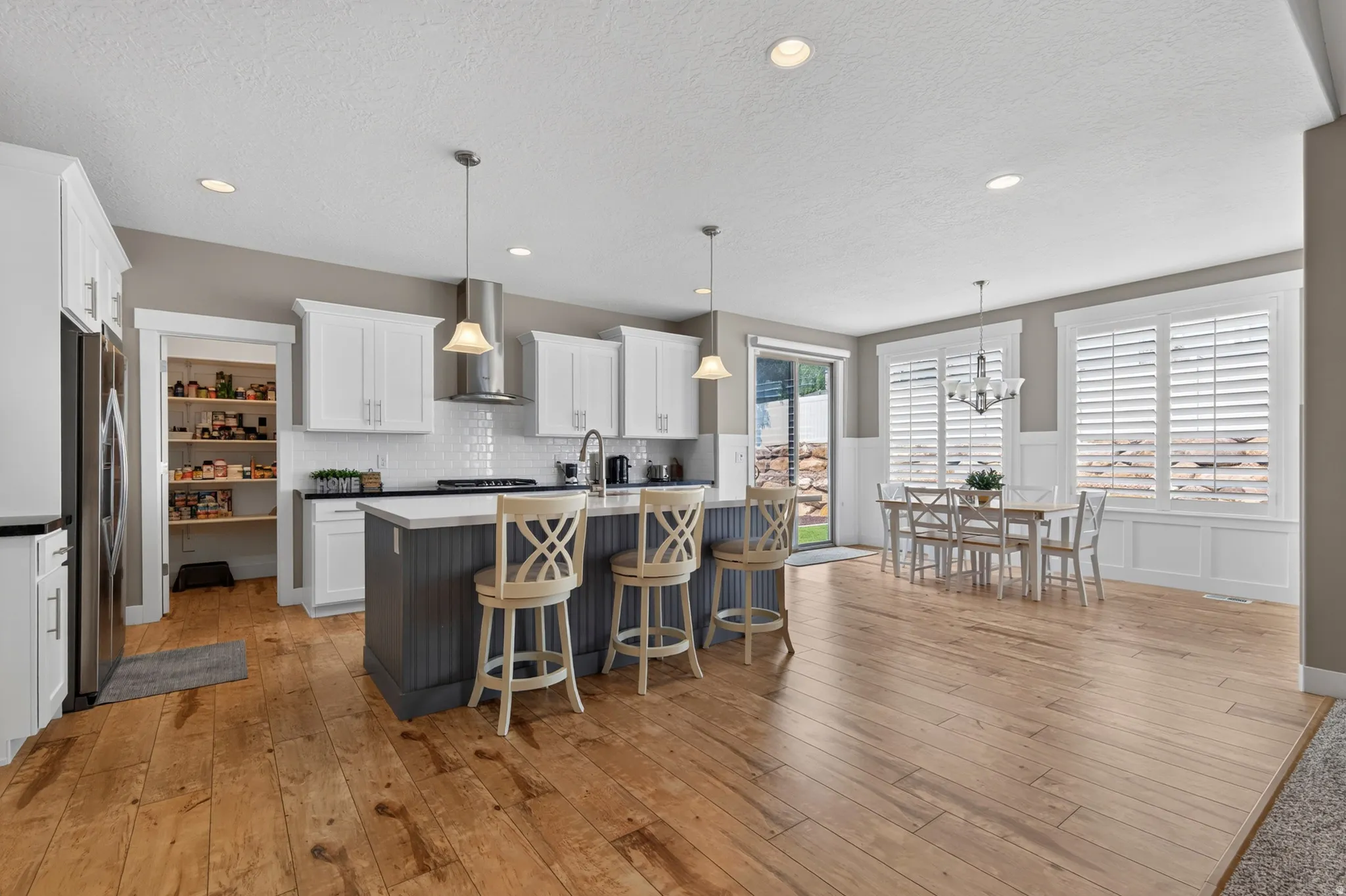 Open-concept kitchen and dining area featuring wide plank wood-finish flooring, white shaker cabinetry, a central island with bar seating, and a walk-in pantry
