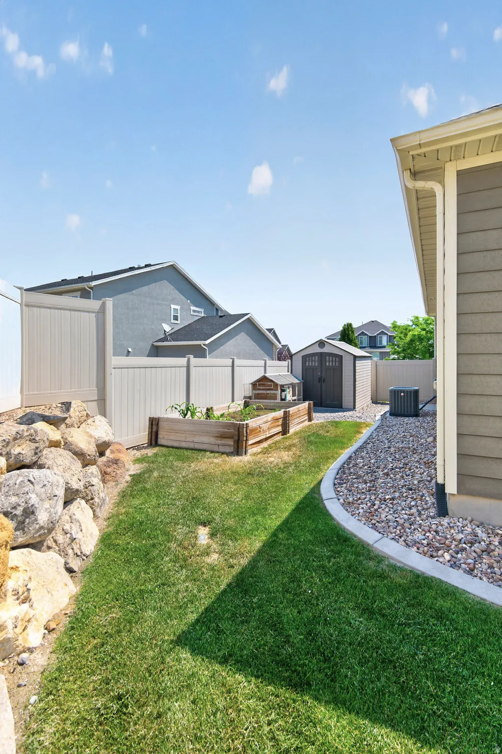 Manicured lawn area featuring a stone retaining wall, raised garden beds, a gravel border, and a durable vinyl fence