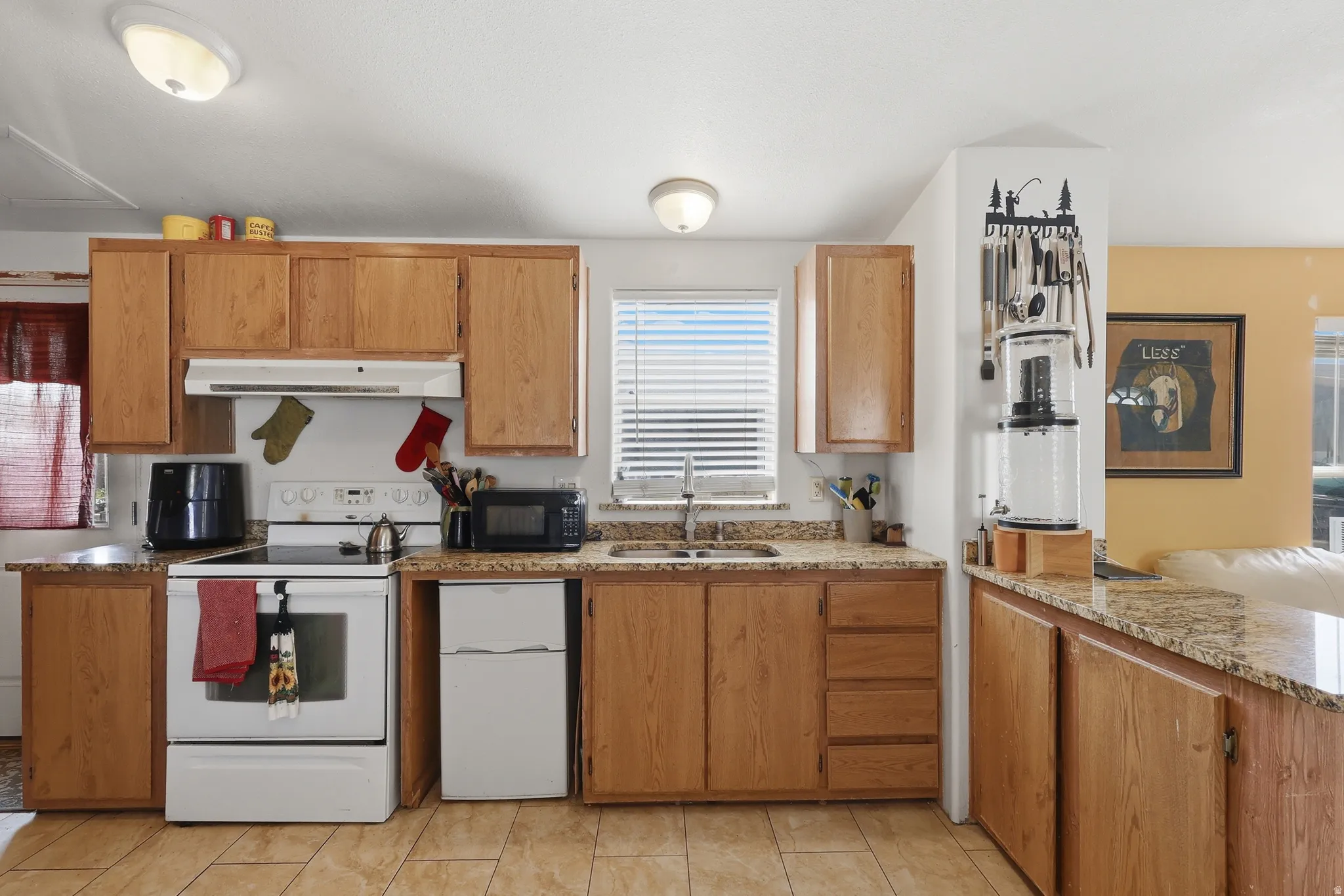 Kitchen featuring wood-finish cabinetry, light-toned granite-style countertops, white electric range with overhead vent, stainless steel sink with window above, and tiled flooring