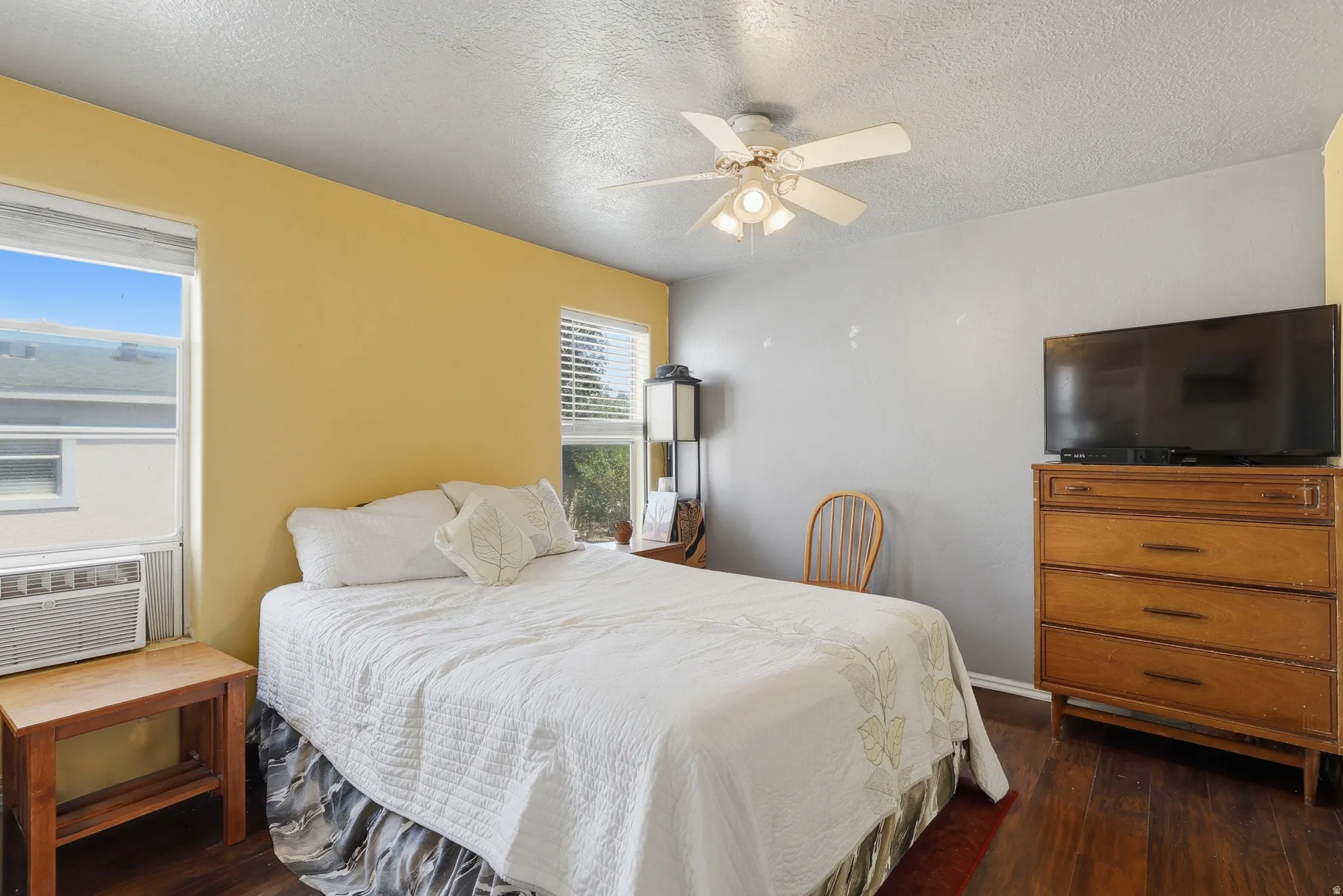 Room featuring wood-finish flooring, multiple windows providing natural light, a ceiling fan with integrated lighting, and an in-wall air conditioning unit