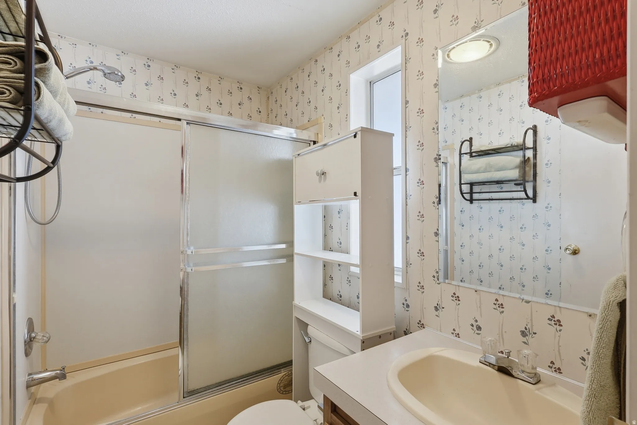 Bathroom featuring a bathtub with a sliding glass enclosure, a vanity with an integrated sink, a built-in wall cabinet with open shelving, a wall-mounted mirror, and a window for natural light