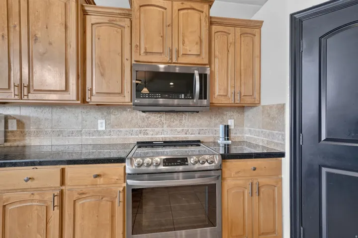 Kitchen featuring light wood cabinetry, stainless steel oven with an electric cooktop, built-in microwave, dark countertops, and a neutral tile backsplash