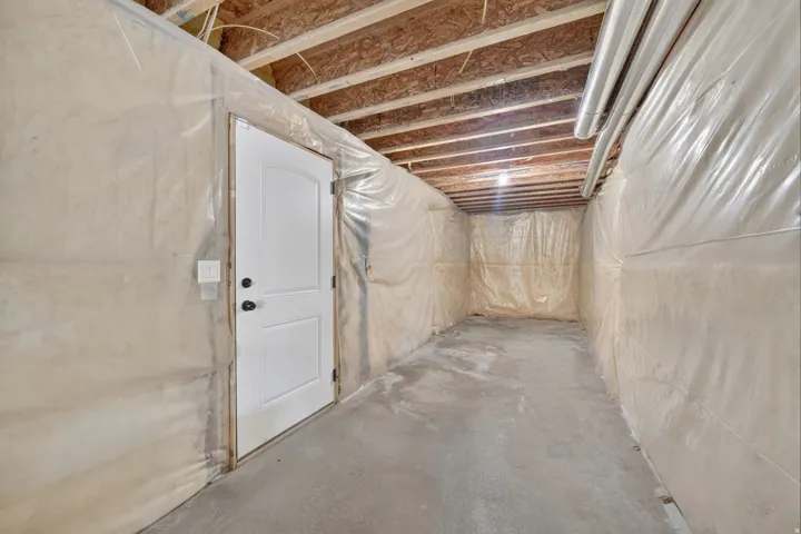 Unfinished basement space featuring a white paneled door with black hardware, exposed ceiling joists, concrete flooring, and translucent wall coverings