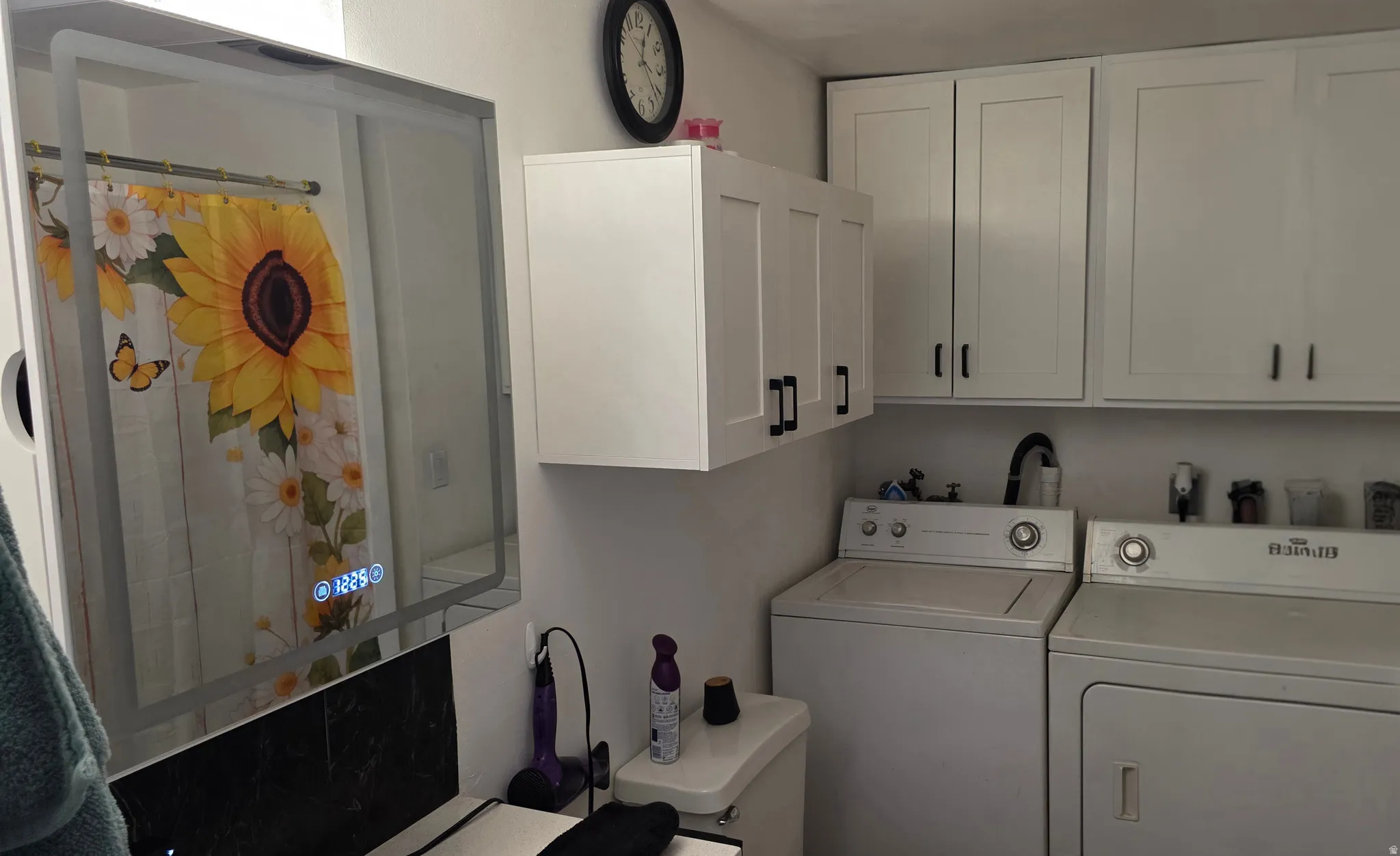 Laundry area featuring white cabinetry with black hardware, side-by-side washer and dryer, and an illuminated vanity mirror
