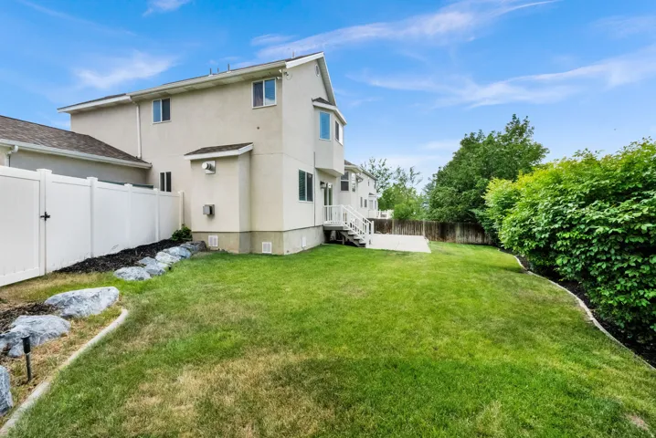 Spacious backyard featuring a lush lawn, mature hedges, a concrete patio, and a white vinyl privacy fence