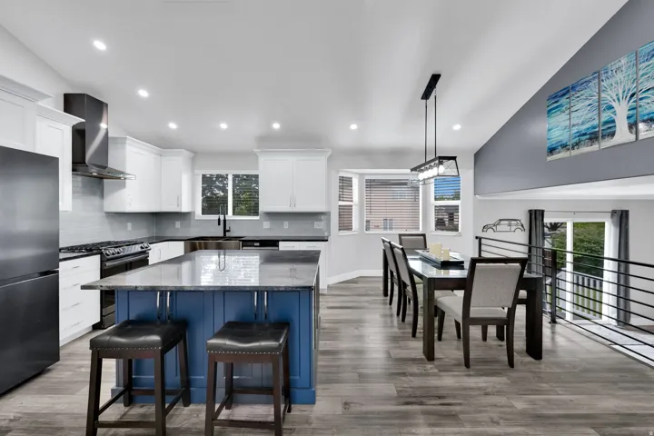 Open-concept kitchen and dining area featuring upgraded NuCore performance ridged-core LVP flooring,, custom white cabinetry, a blue kitchen island with seating, a black stainless steel range hood, and a dining area with a modern chandelier and bay window