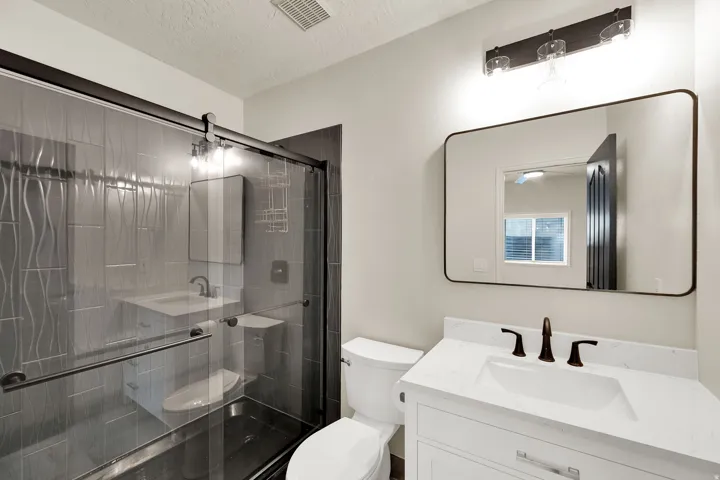 Bathroom featuring a glass-enclosed shower with textured tile, white vanity with an integrated sink, dark bronze fixtures, and a rectangular framed mirror