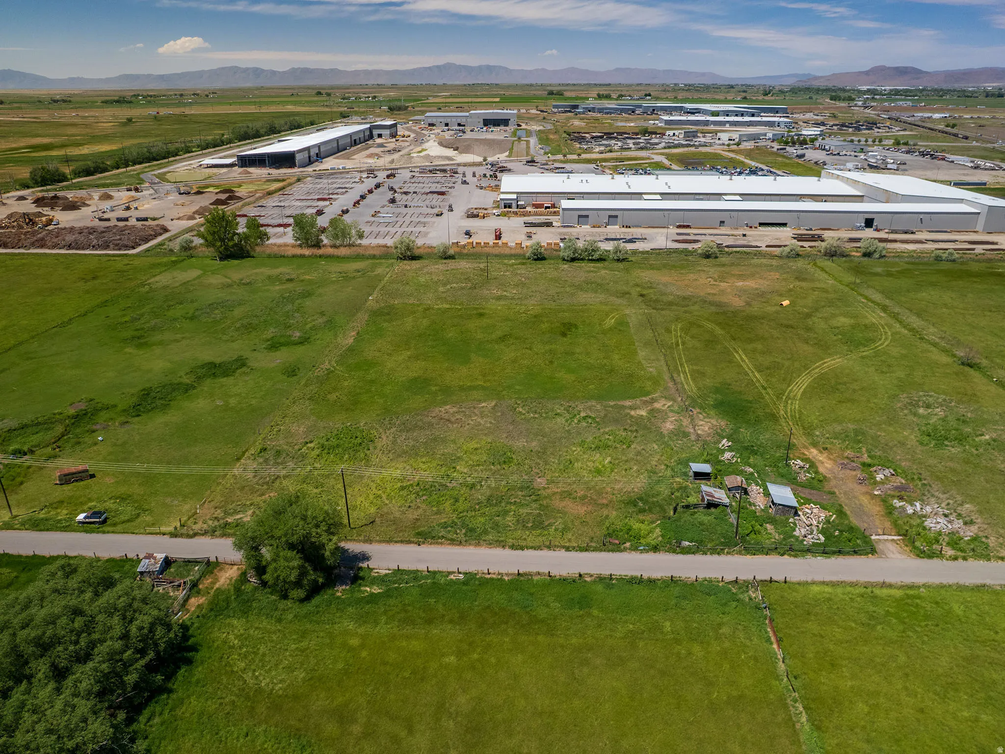 Expansive green fields featuring a paved road, scattered trees, and distant industrial structures under a clear sky with mountains on the horizon