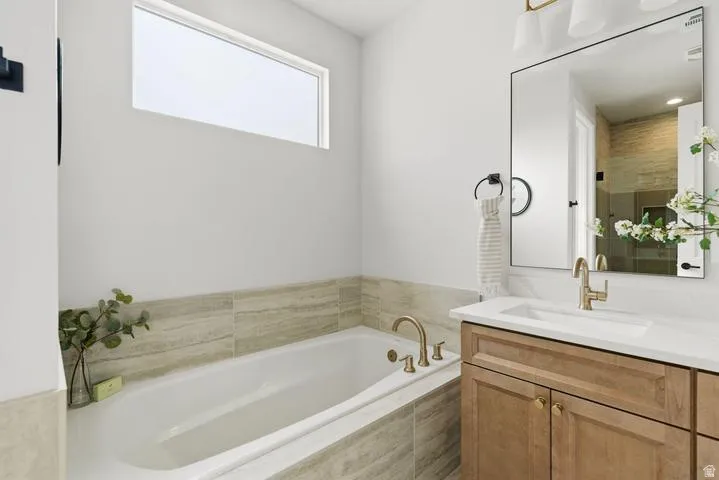 Built-in soaking tub with light-toned tile surround, single vanity with wood-finish cabinetry, white countertop, and gold-finish hardware