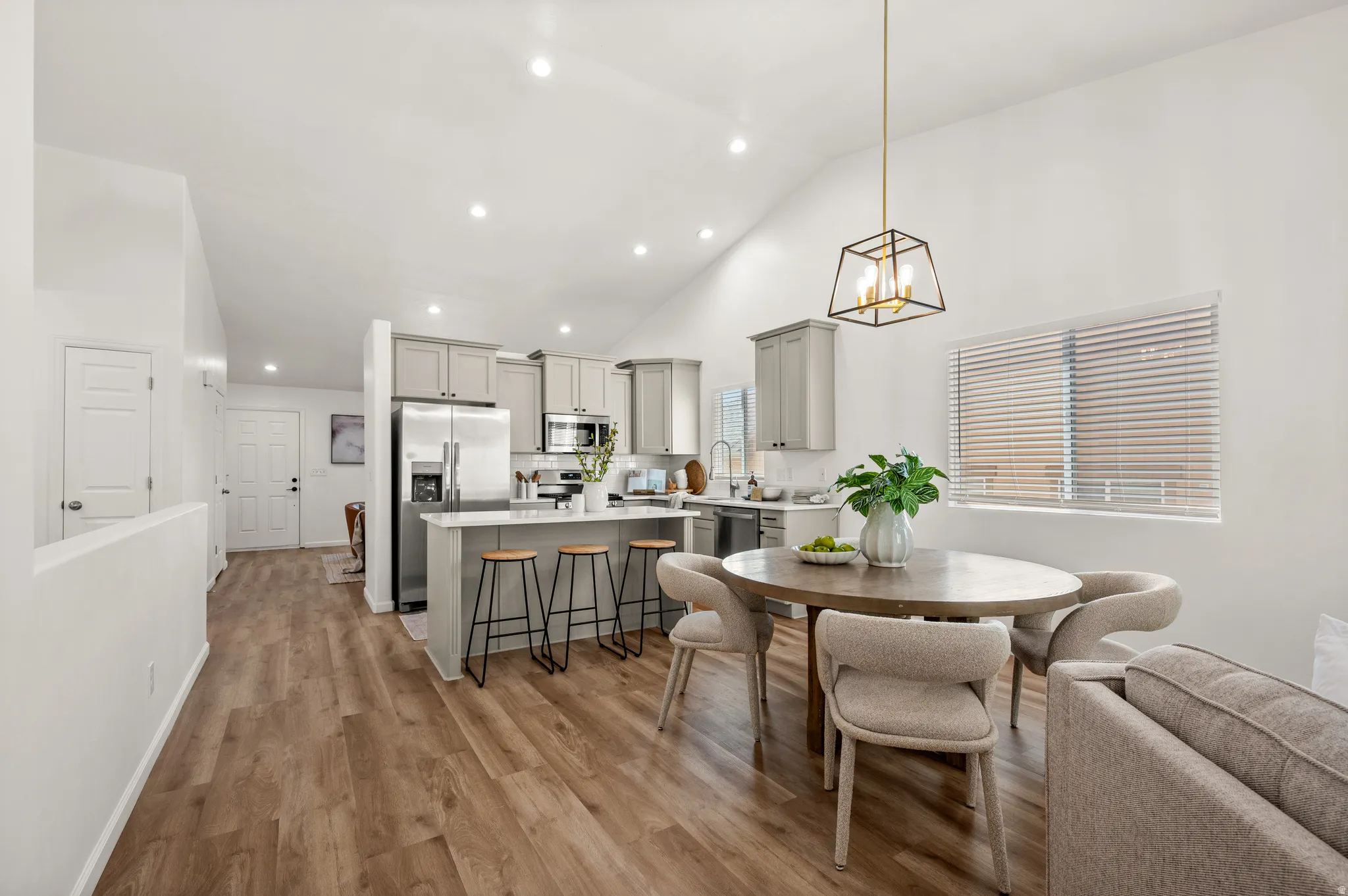 Vaulted ceilings enhance this open-concept space featuring wood-finish flooring, a contemporary kitchen with light gray cabinetry and stainless steel appliances, recessed lighting, and a modern dining pendant light fixture