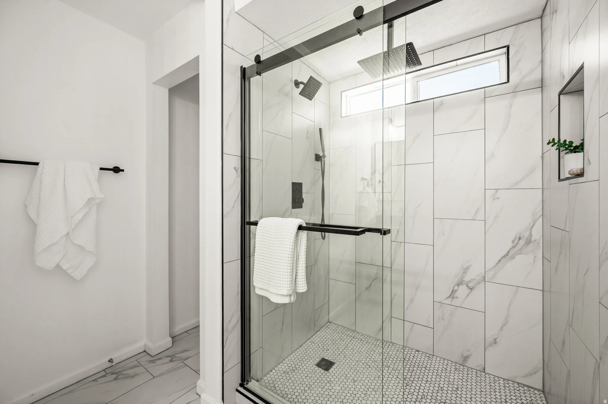 Walk-in shower featuring a frameless glass enclosure with matte black hardware, white marbled-pattern wall tiling, penny tile flooring, a horizontal transom window, and a built-in wall niche