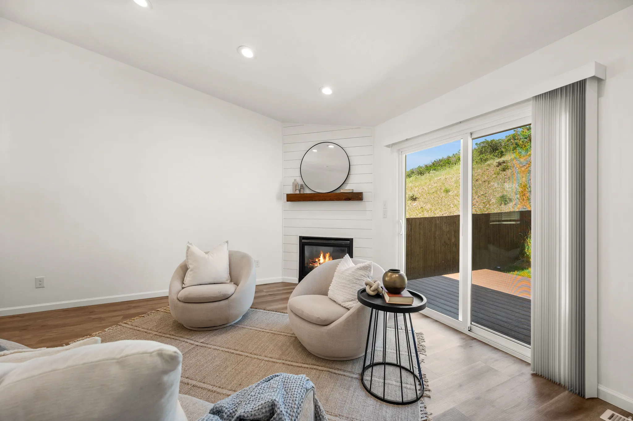 Living area featuring wood-finish flooring, a corner shiplap-accented fireplace with a wood mantel, recessed lighting, and a sliding glass door opening to an outdoor deck
