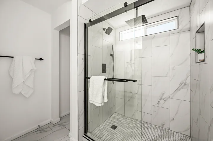 Walk-in shower featuring a frameless glass enclosure with matte black hardware, white marbled-pattern wall tiling, penny tile flooring, a horizontal transom window, and a built-in wall niche