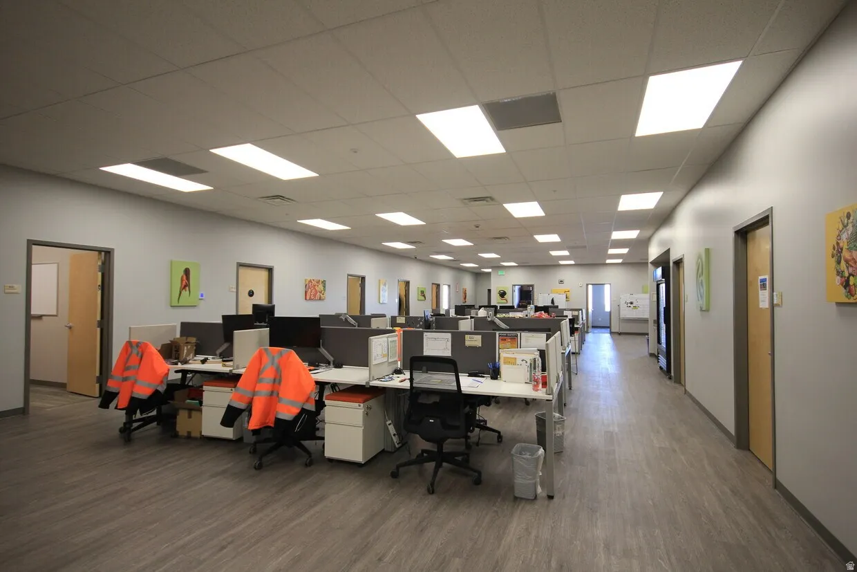 Open-plan office area featuring light wood-finish flooring, drop ceiling with recessed lighting, and light grey painted walls