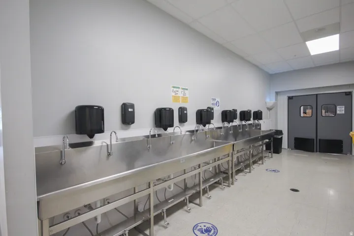 Stainless steel trough sinks with multiple chrome faucets, wall-mounted soap dispensers, and a light gray wall finish