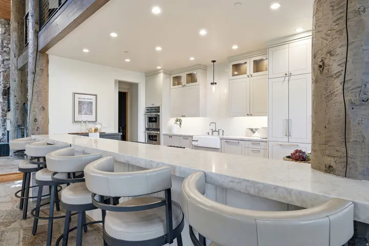 Kitchen featuring an extended island with a light-toned countertop, white cabinetry with glass upper doors, a farmhouse sink, and integrated double ovens
