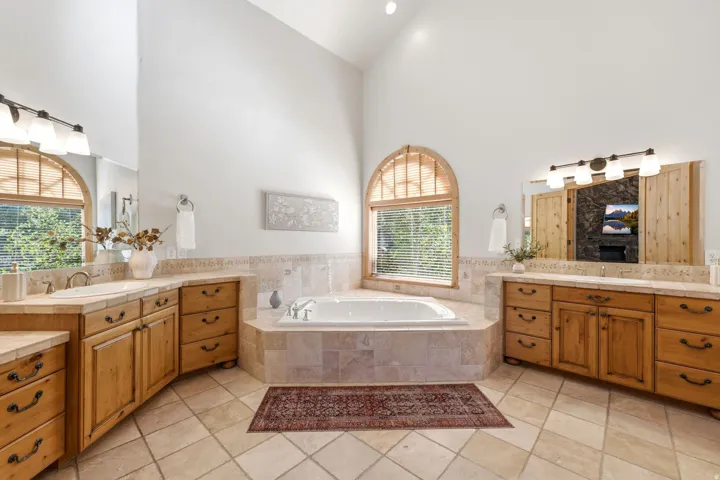 Spacious bathroom featuring a corner soaking tub, arched window with wood blinds, double vanity with wood cabinetry, tile flooring, and vaulted ceiling