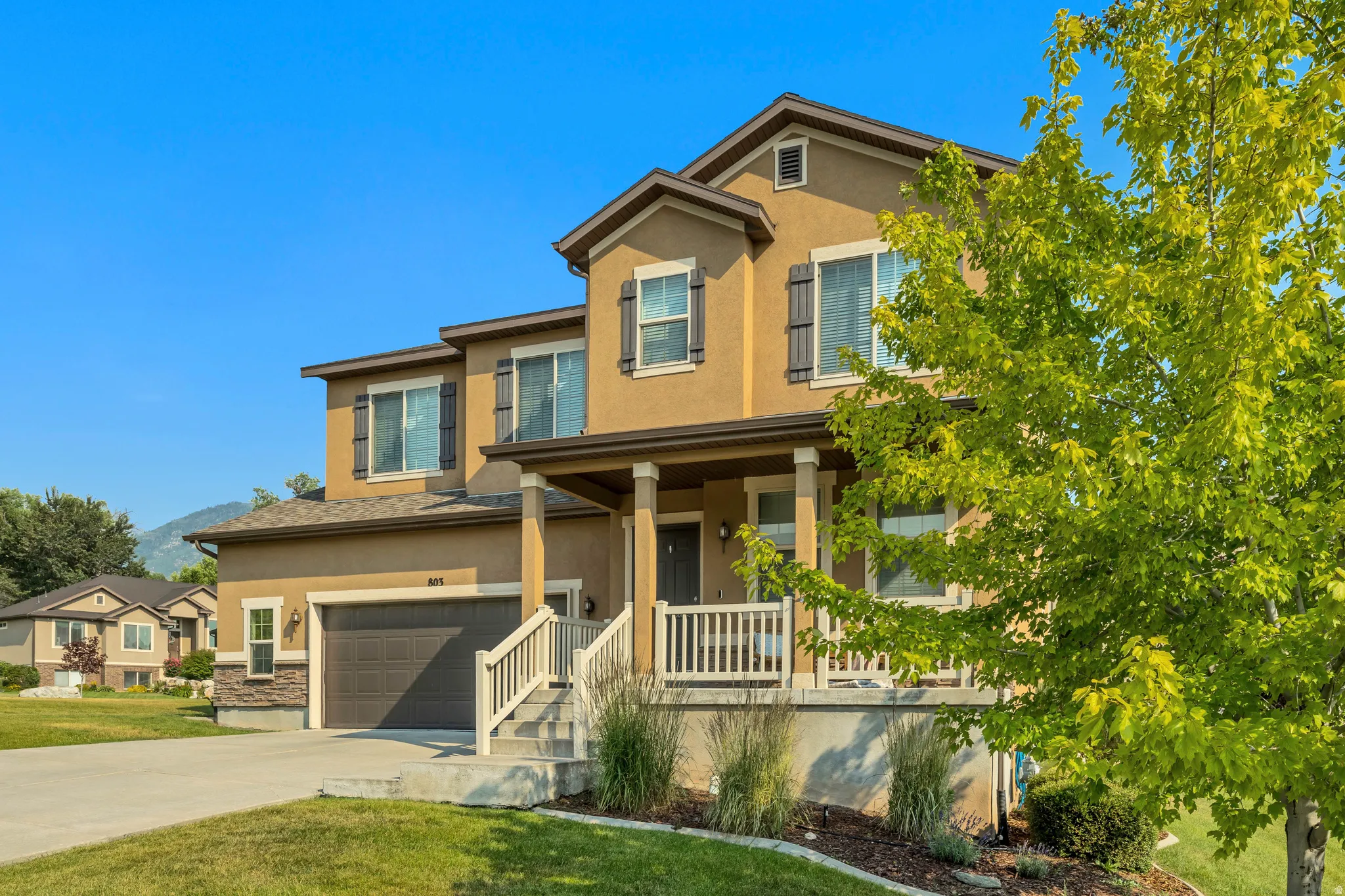 Contemporary two-story residence featuring a stucco exterior with stone accents, a covered front porch, an attached garage, a mature tree, and a landscaped front yard