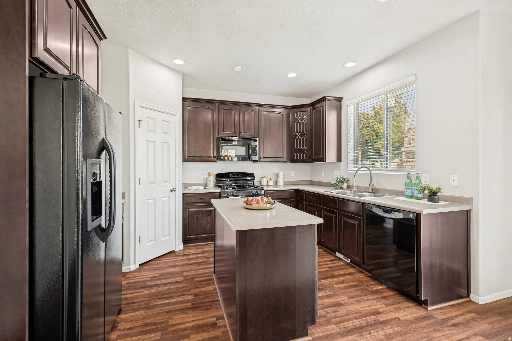 Kitchen featuring wood-finish flooring, dark wood cabinetry, a central island with a light-toned countertop, recessed lighting, and a window with blinds