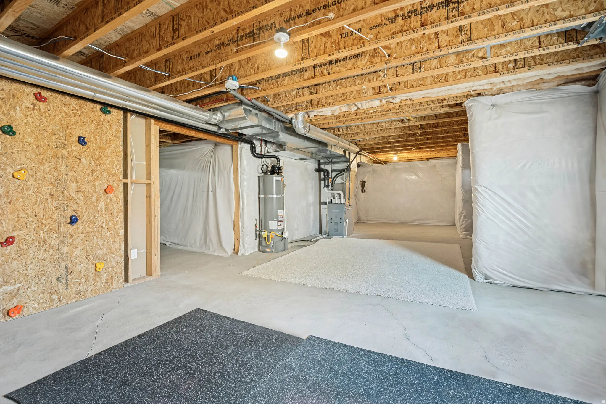 Unfinished basement featuring exposed wood joists, concrete flooring, utility access, a water heater, and a pre-framed wall section with climbing holds