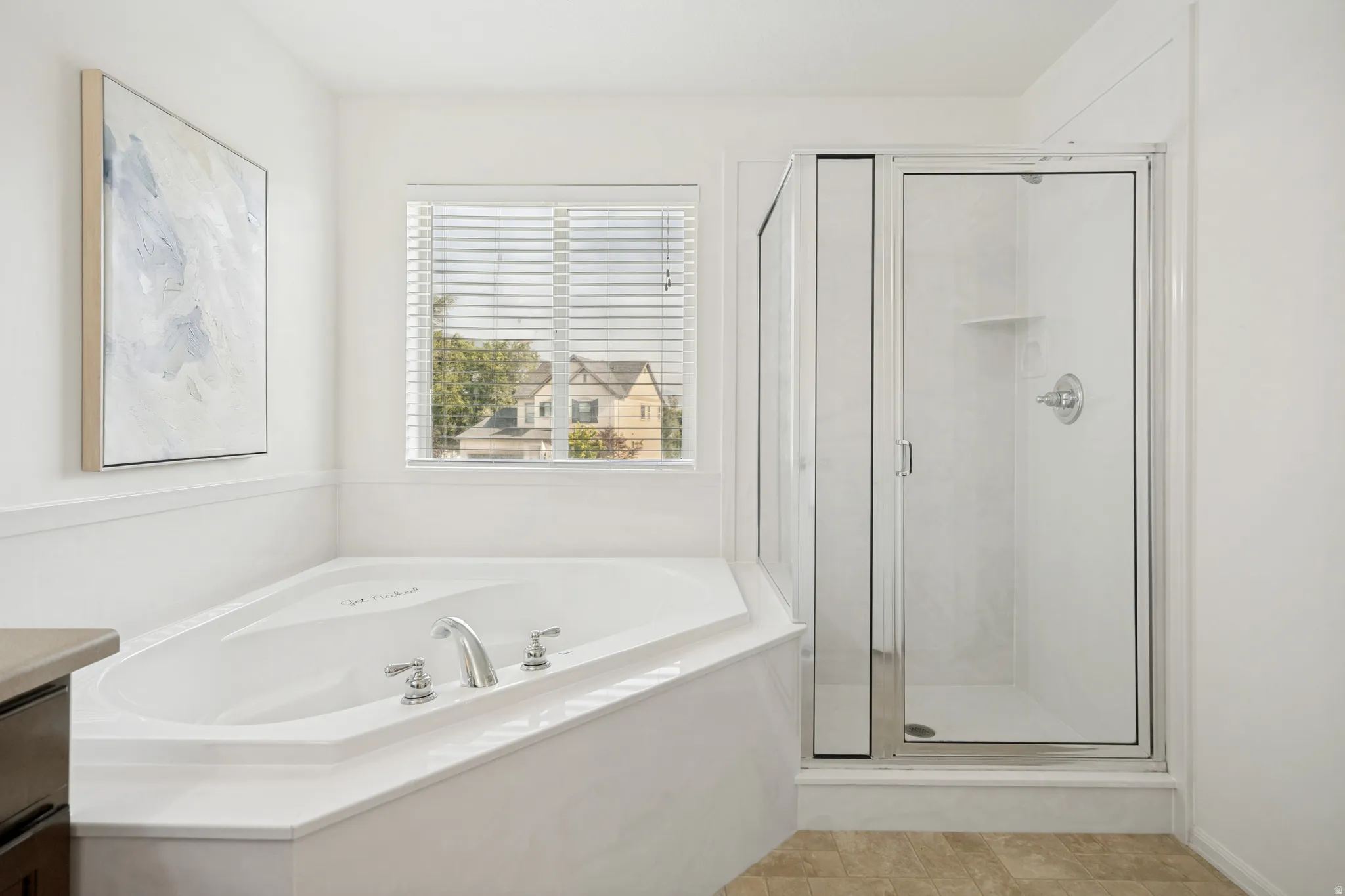 White corner soaking tub with chrome fixtures, glass-enclosed shower, white wall paneling, and a window with horizontal blinds