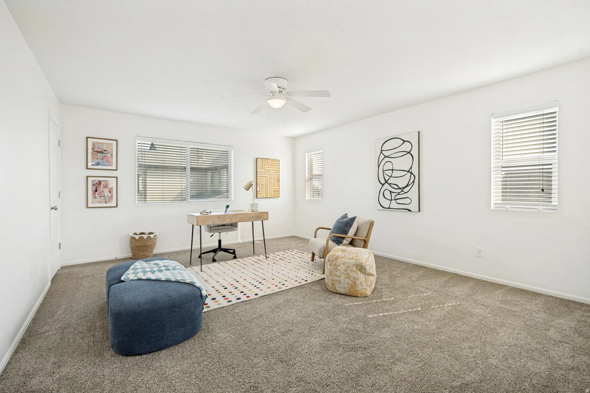 Spacious room featuring neutral carpet flooring, multiple windows with blinds, a ceiling fan, and white painted walls