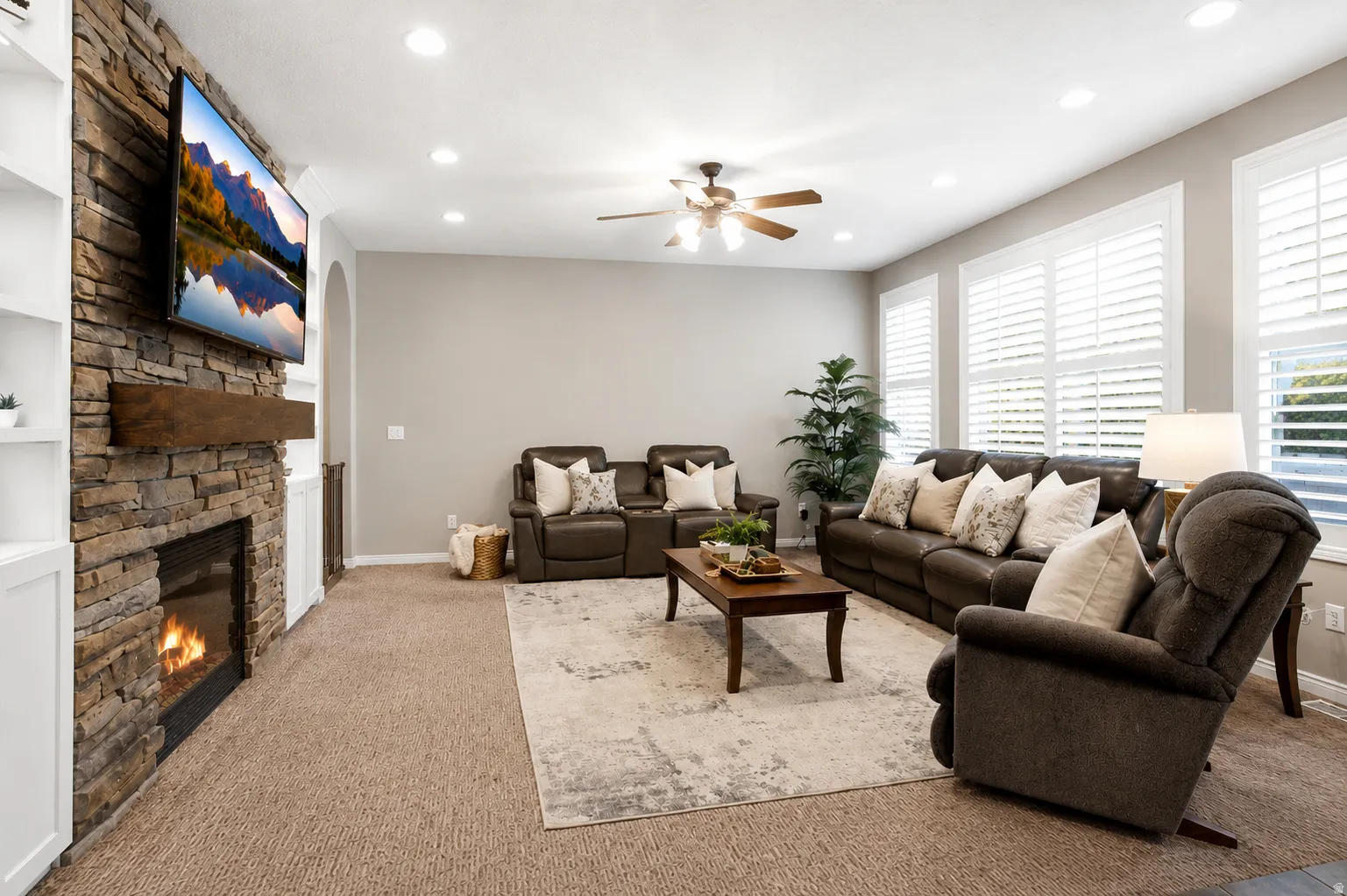Spacious living area featuring a stone-clad fireplace with a wood mantle, built-in shelving, recessed lighting, and multiple windows with plantation shutters