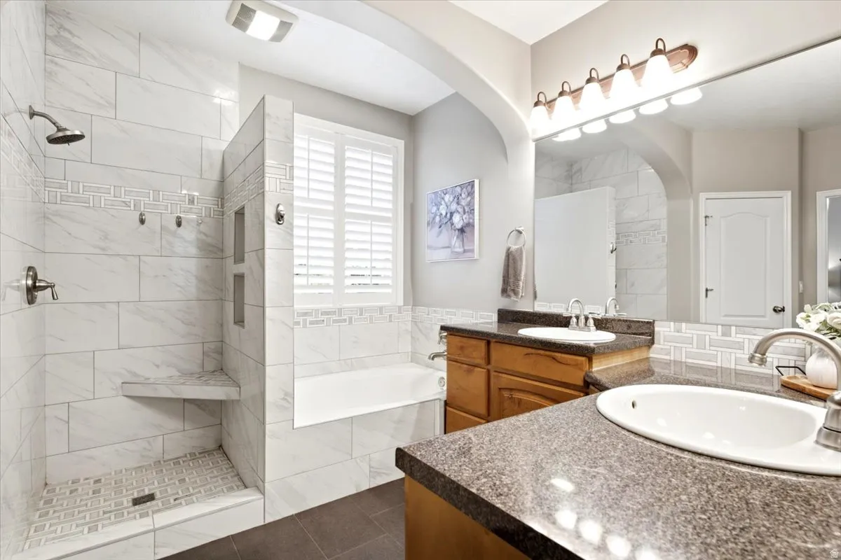 Full bathroom featuring a walk-in shower with a built-in bench and tiled niche, a separate bathtub with plantation shutters, a dual vanity with undermount sinks and dark stone-finish countertops, and a large wall-mounted mirror