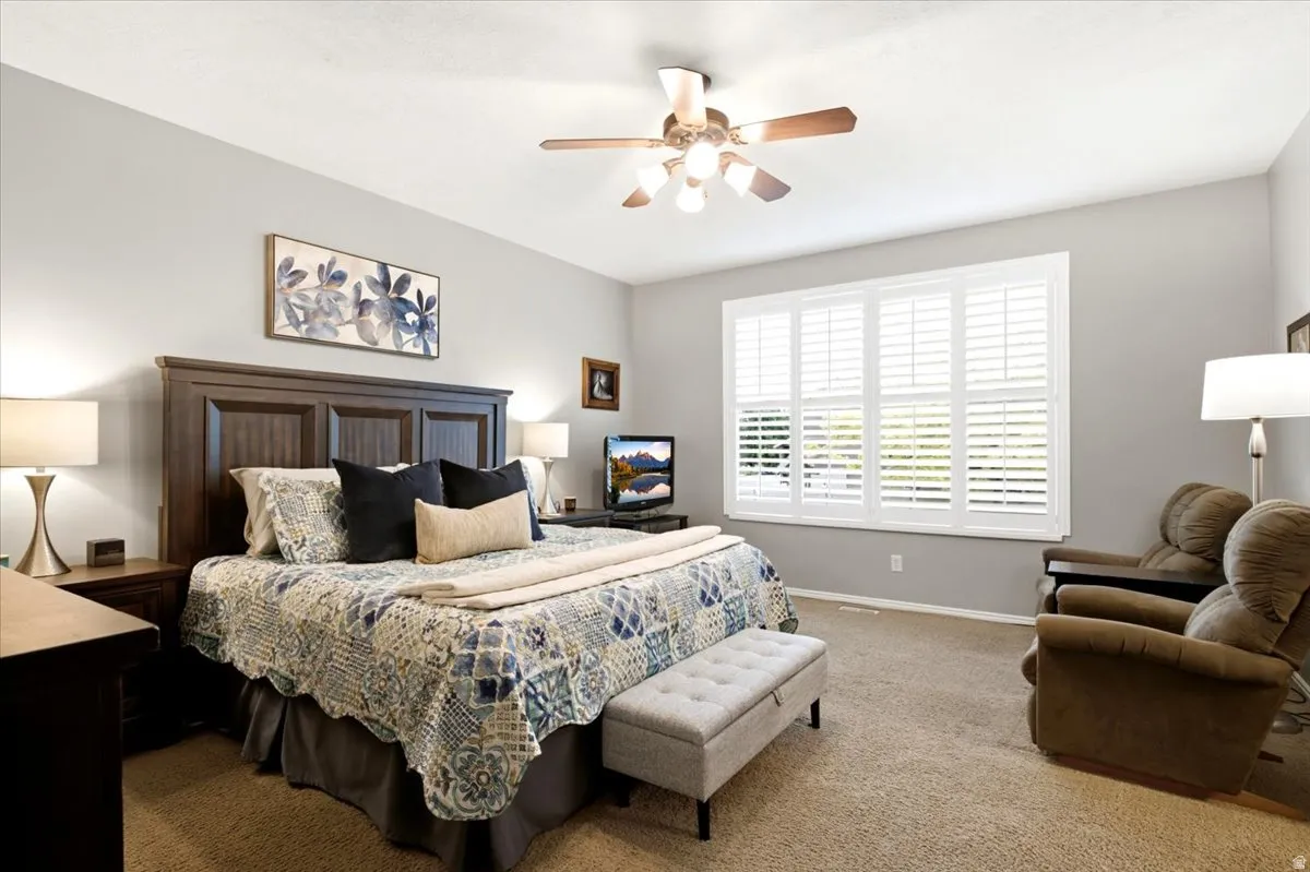 Spacious room featuring a large window with white plantation shutters, neutral-toned carpeting, and a ceiling fan with integrated lighting