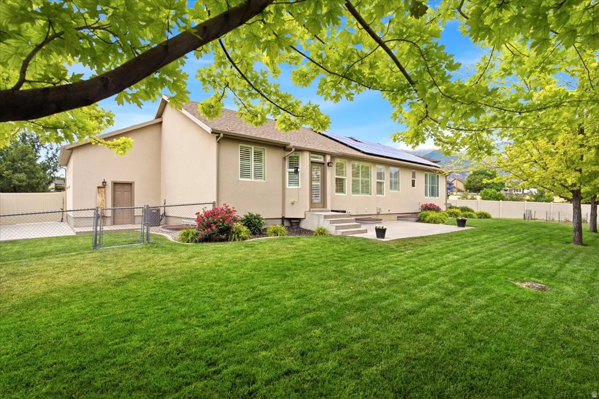 Expansive rear yard featuring a concrete patio, established lawn, mature trees, and solar panels on the roof