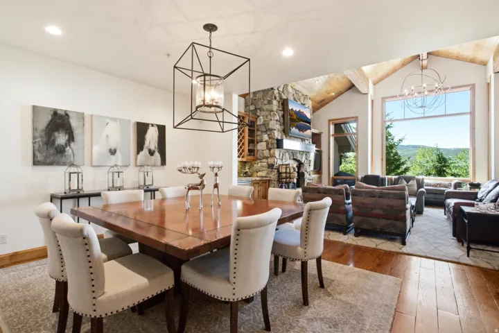 Open-concept living space with vaulted ceilings featuring exposed wood beams, a stone-clad fireplace, and large picture windows