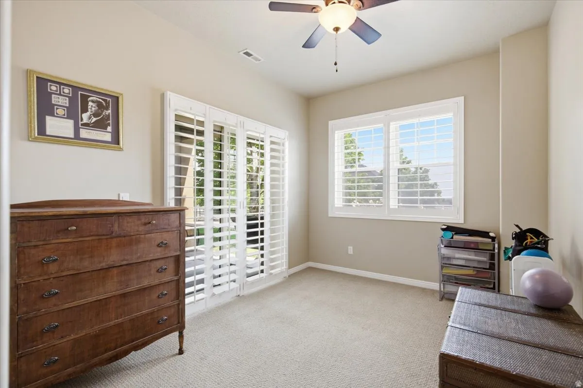 Carpeted interior space featuring a ceiling fan with integrated lighting, a glass sliding door with plantation shutters, and a window with plantation shutters
