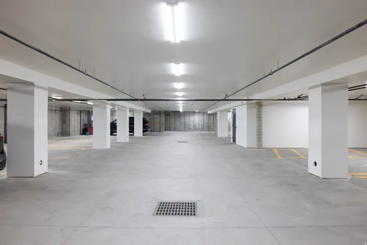 Spacious underground parking garage featuring concrete flooring, exposed concrete walls, white structural columns, and overhead fluorescent lighting