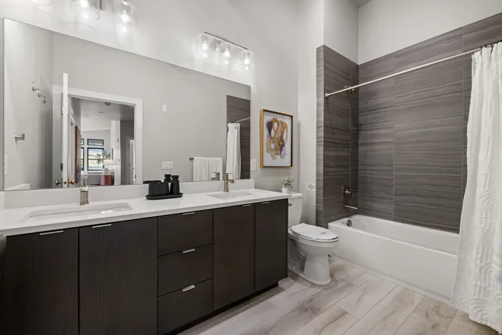 Bathroom featuring a dual vanity with undermount sinks and individual faucets, a full-width mirror, and a combination shower-tub with dark grey wall tiling