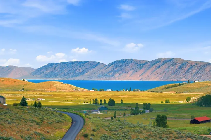 Expansive landscape featuring a winding asphalt road, verdant hillsides, distant blue lake, and mountain range backdrop
