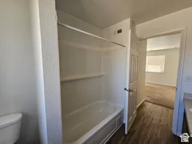 Bathroom featuring a white tub/shower combination with integrated shelving, a white toilet, and wood-finish flooring
