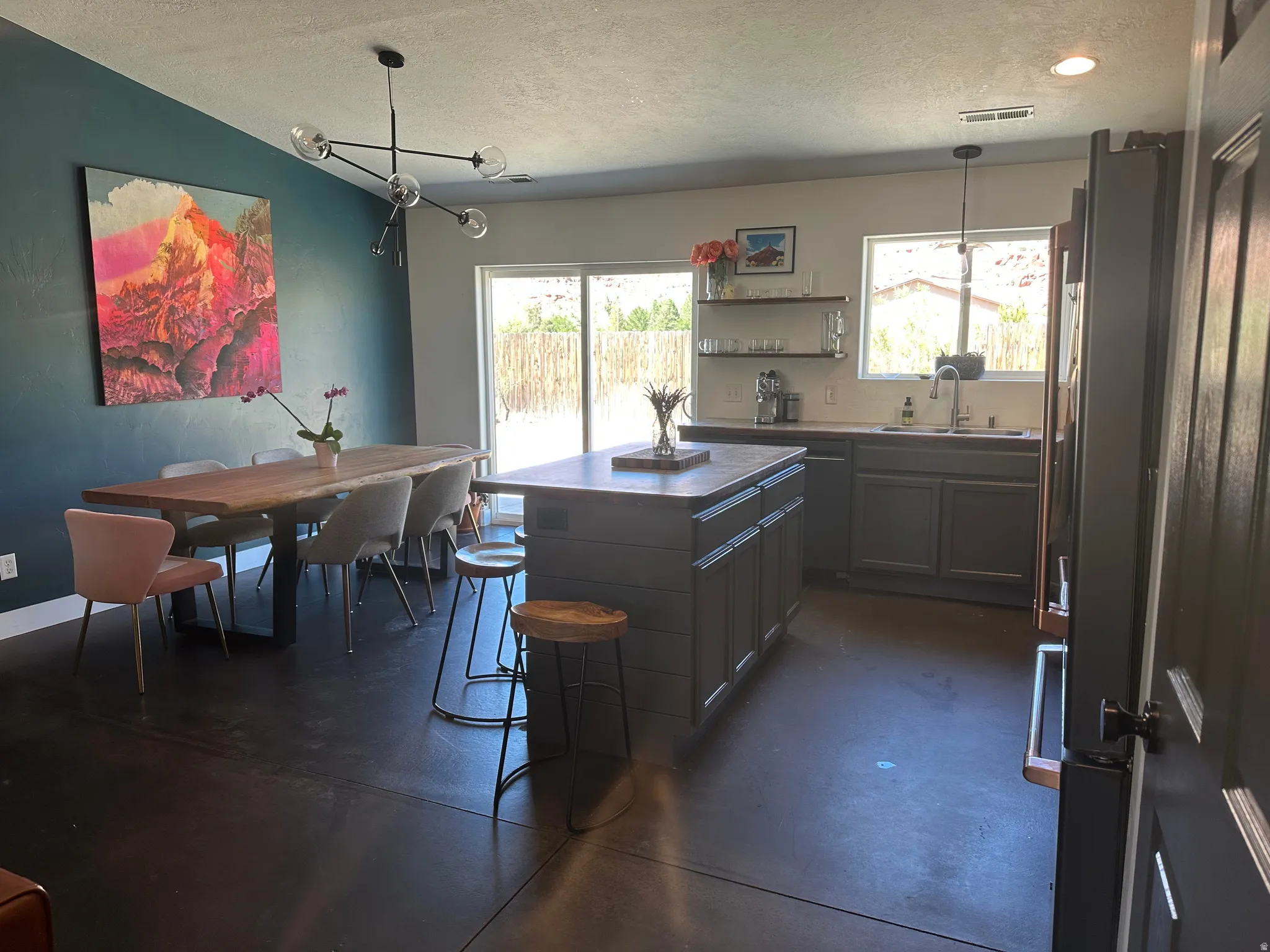 Open-concept kitchen/dining space featuring stained concrete flooring, a kitchen island with dark shaker-style cabinetry, and a sliding glass door providing exterior access