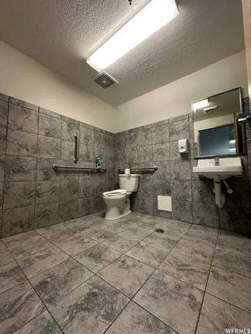 Restroom featuring floor-to-ceiling gray patterned tile, wall-mounted sink, toilet, and grab bars