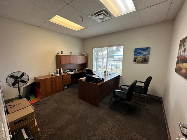 Commercial office space featuring textured carpet flooring, dropped acoustic ceiling tiles, recessed fluorescent lighting, and a window with horizontal blinds