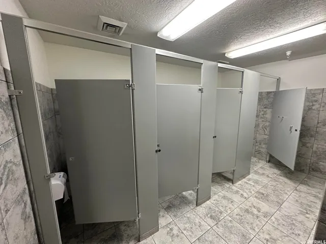 Commercial restroom featuring multiple stalls with privacy partitions, grey tile flooring, and a coordinating grey-tiled wainscoting