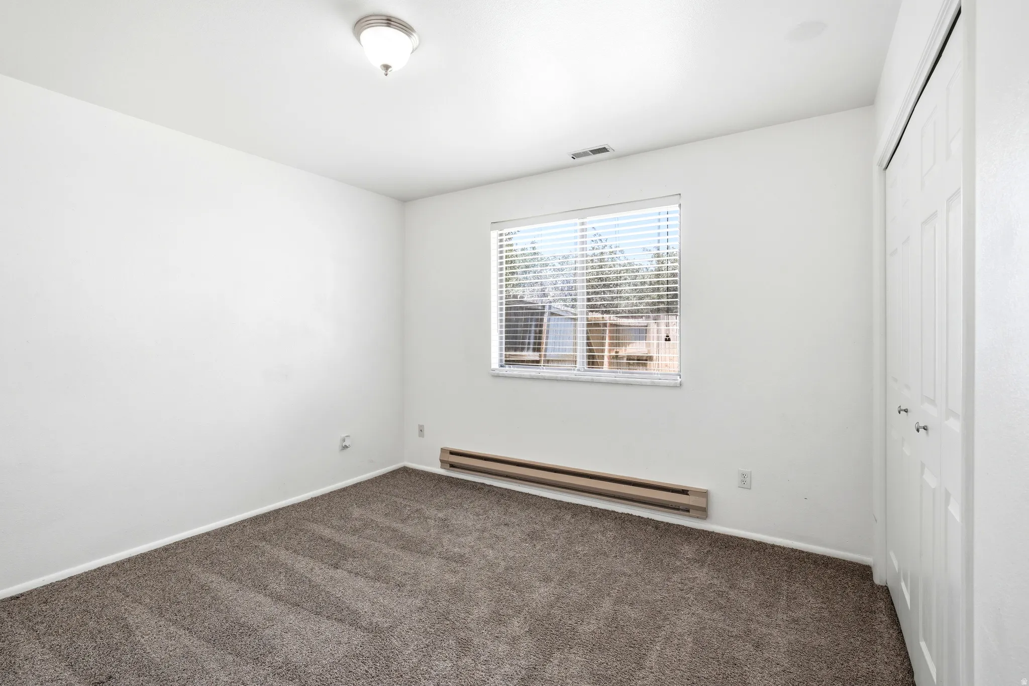 Spacious interior room featuring a neutral color palette, plush carpet flooring, a window with horizontal blinds, built-in bi-fold closet doors, and a ceiling-mounted light fixture