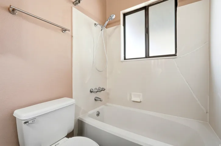 Bathroom featuring a white tub-shower combination with a showerhead and separate faucet, a window with frosted panes, and a white toilet