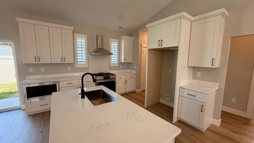 Kitchen featuring white shaker-style cabinetry, a large kitchen island with an undermount sink, gas range with stainless steel hood, quartz countertops, and wood-finish flooring