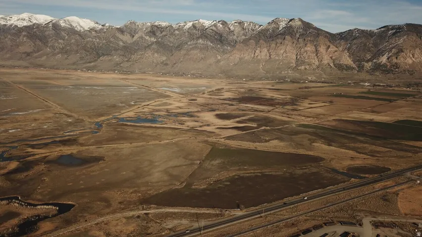 Expansive high desert landscape featuring a prominent mountain range with snow-capped peaks, and a highway traversing the foreground