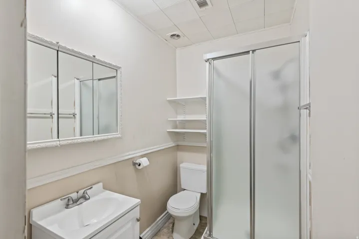 Bathroom featuring a white vanity with integrated sink, a framed medicine cabinet, a toilet, a shower with frosted glass enclosure, and wall-mounted shelving
